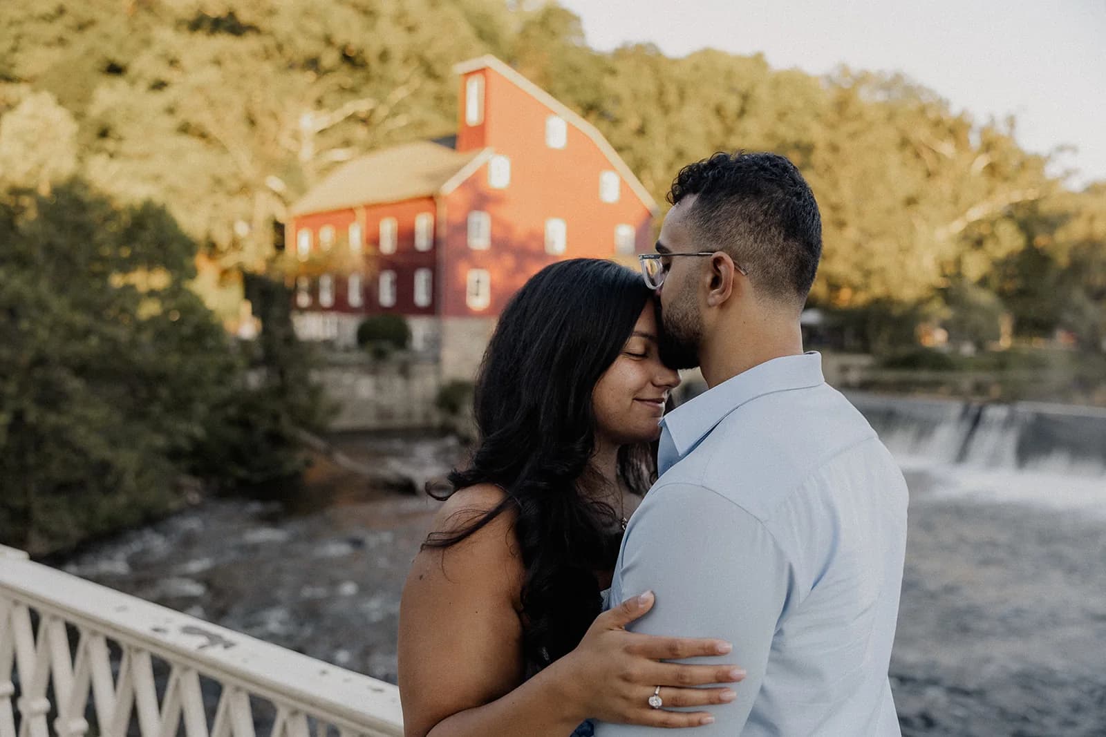 Anthony kissing Lily on the forehead on a bridge in front of the Red Mill